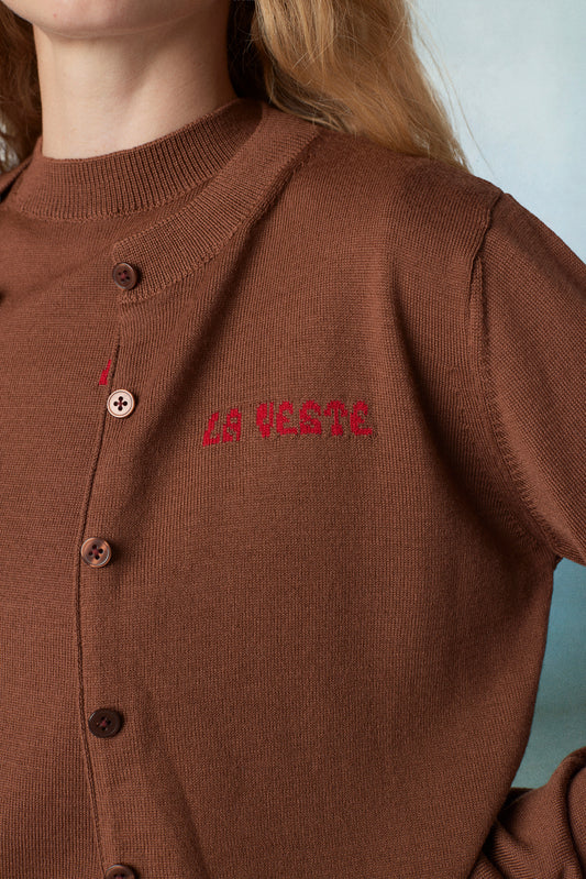 Close-up of a person wearing a brown ribbed knit cardigan sweater with brown buttons and red embroidered text reading 'LA VESTE' on the chest.