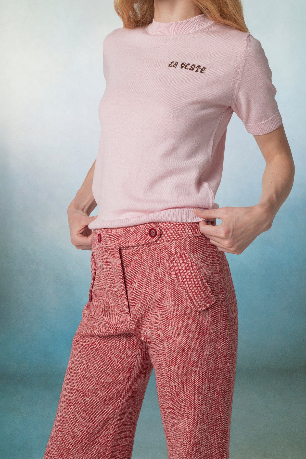 A model wearing a light pink short-sleeve knit top with 'LA VESTE' embroidered on the chest, paired with textured dusty red trousers, set against a blue gradient studio background.